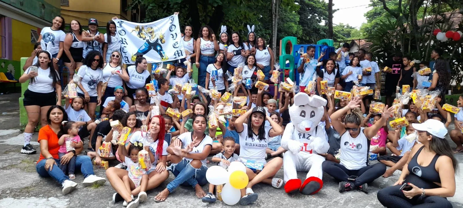 Um grande grupo de pessoas está sentado no chão com balões e um mascote.