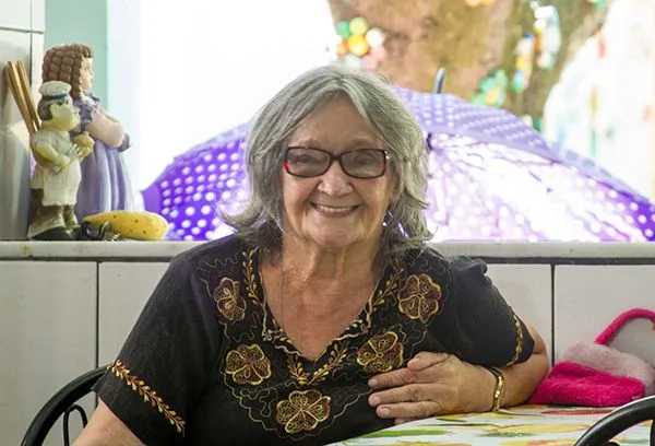 Uma senhora idosa usando óculos está sentada em uma mesa sorrindo.